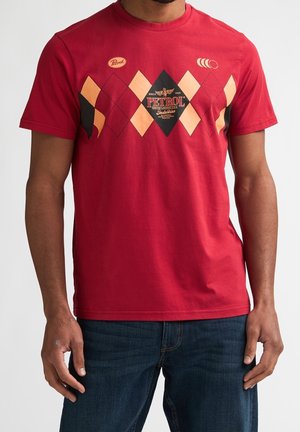 Mand iført rød kortærmet t-shirt med diamantmønster og "Petrol"-logo, parret med mørkeblå jeans, vist fra hals til hofter.
