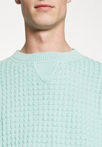 Hellblauer, gestrickter Pullover mit strukturiertem Muster, versehen mit einem gerippten Rundhalsausschnitt und einem kleinen V-förmigen Detail am Kragen.