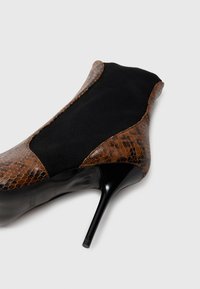 Brun slangeskinn og svart stoff ankelstøvlett med spiss tå og høy stiletthæl på hvit bakgrunn.