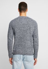 Man som bär en långärmad, grå och vit melerad tröja och ljusblå jeans, sedd bakifrån mot en neutral bakgrund.