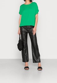 Haut vert à manches courtes avec un col rond, associé à un pantalon large en cuir noir. Tenant un sac à main tissé noir et portant des sandales à glissement noires.