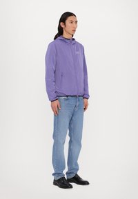 Casaco roxo com fecho éclair, apresentando um capuz e bolsos frontais, combinado com jeans azul claros e sapatos pretos, em pé sobre um fundo neutro.