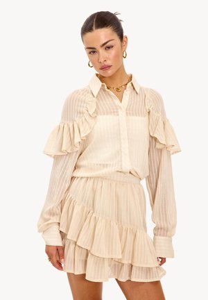 Femme portant un chemisier transparent crème à volants et une jupe à volants assortie à étages, avec des boucles d'oreilles créoles dorées et un collier, posant devant un fond blanc.