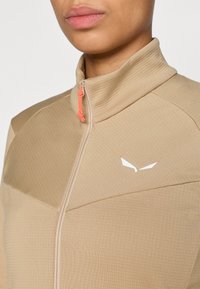 Μπεζ ζακέτα με φερμουάρ και ψηλό γιακά, υφασμάτινη υφή και λευκή λογότυπο λεπτομέρεια. Διαθέτει ένα φωτ εινό πορτοκαλί τράβηγμα φερμουάρ.