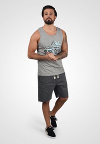 Camisa sin mangas gris con un gráfico y texto de temática playera, combinada con pantalones cortos oscuros. Zapatillas negras con suelas blancas. Atuendo casual.
