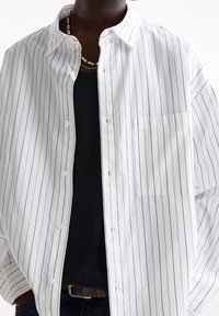 Personne portant une chemise blanche à fines rayures verticales noires, ouverte sur un haut noir, avec un collier de perles visible au niveau du cou.