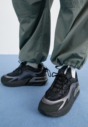 Personne portant des baskets noires et grises avec des semelles épaisses et un pantalon cargo vert debout sur un tapis bleu clair.
