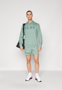 Sudadera deportiva verde claro y pantalones cortos con el logo "PUMA", combinados con zapatillas blancas. Modelo sosteniendo un bolso negro.