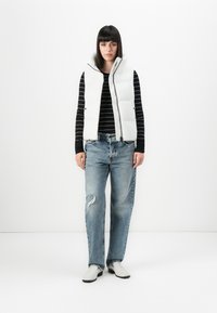 Vrouw met steil, donker haar, gekleed in een witte puffervest, een zwart gestreept langetermijnshirt, afgescheurde blauwe jeans en witte schoenen, staand tegen een witte achtergrond.