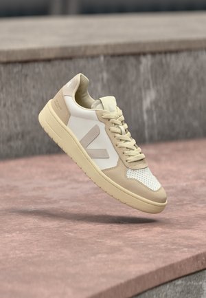 Baskets beige et blanches avec une semelle épaisse et des lacets, flottant au-dessus d'une surface en pierre avec un mur en pierre gris flou en arrière-plan.