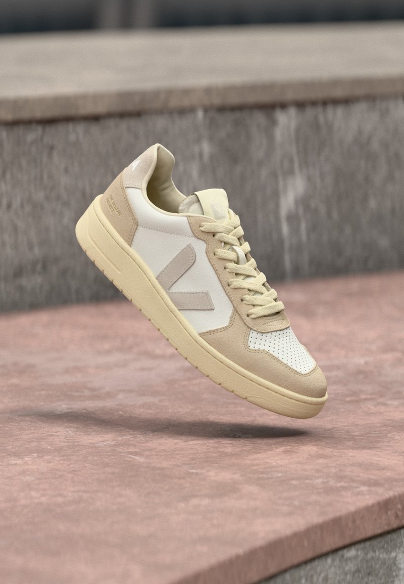 Μπεζ και λευκό αθλητικό παπούτσι με χοντρή σόλα και κορδόνια, που αιωρείται πάνω από μια πέτρινη επιφάνεια με θολό γκρι πέτρινο τοίχο στο φόντο.