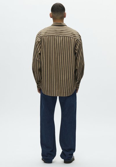 Mężczyzna stojący tyłem, ubrany w brązowo-beżową koszulę w pionowe paski z długim rękawem oraz ciemnoniebieskie jeansy, na białym tle.