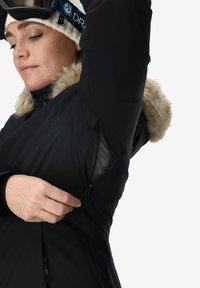 Mujer con chaqueta de invierno negra con capucha de piel abriendo una abertura de ventilación de malla debajo de su brazo levantado.