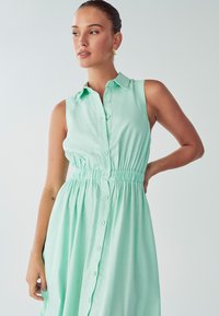 Šviesiai žalia be rankovių suknelė su mygtukų uždarymu priekyje, įtempta juosmens dalimi ir surinktu sijonu. Pagaminta iš lengvo audinio su lygiu paviršiumi.