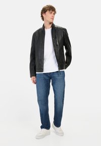Zwarte leren jas met gestikte schouderdetails, voorzame rits, gedragen over een wit shirt, gecombineerd met blauwe jeans en witte sneakers.