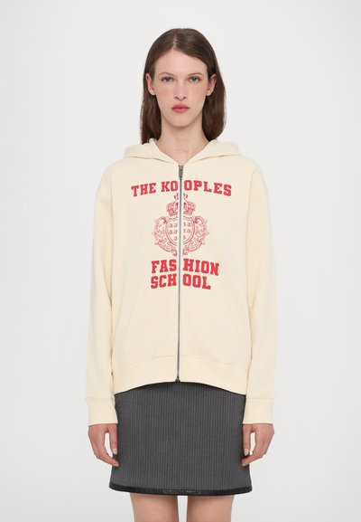 Kermansin zip-up huppari, jossa on punainen graafinen teksti "THE KOPOLES FASHION SCHOOL." Varustettu hupulla ja etutaskuilla, yhdistettynä harmaaseen ribbihameeseen.