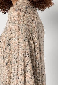 Strickjacke in Beige mit gesprenkelten Mustern. Verfügt über ein Zopfmuster-Design, lockere Passform und weichen Stoff. Sichtbare Nähte und fallender Stoff.