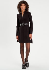 Black zip-up dress with long sleeves, cinched waist by a round-stud belt, paired with black loafers featuring a chain accent.