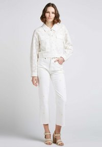 Veste blanche courte avec texture florale et deux poches avant, associée à un jean blanc avec broderie florale jaune et des sandales à plateforme beiges.
