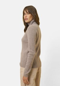 Donna con capelli castani che indossa un maglione beige a collo alto e pantaloni color beige chiaro, in profilo su uno sfondo bianco uniforme.