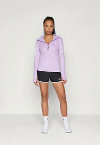 Lys lilla treningsjakke med glidelås og lange ermer, med tre hvite striper på venstre arm, kombinert med svarte shorts og hvite sokker.