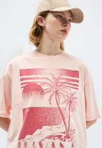 Νέος άνθρωπος που φοράει μπεζ καπέλο και ροζ ανοιχτό μπλουζάκι με κόκκινο τροπικό σχέδιο με φοινικόδεντρα και δύση του ηλίου.