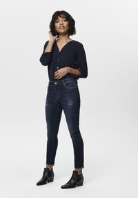 Blusa marina con scollo a V, dettagli con bottoni, abbinata a jeans in denim scuro e stivaletti neri con borchie. Sfondo da studio.