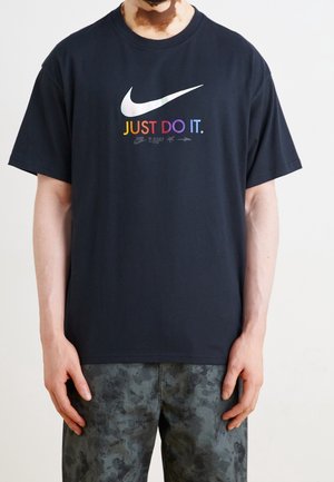 Person iført en sort Nike t-shirt med en flerfarvet "JUST DO IT." slogan og sølv swoosh logo, kombineret med grå mønstrede bukser.
