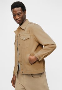 Άνδρας που φοράει μπεζ μπουφάν με κέντημα λουλουδιού και τη λέξη «marimekko», τα χέρια στις τσέπες, στέκεται μπροστά σε λευκό απλό φόντο.