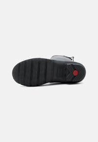 Σόλα μαύρης λαστιχένιας μπότες με υφή λαβής, οριζόντιες γραμμές πέλματος και κόκκινο κυκλικό λογότυπο κοντά στη φτέρνα σε απλό λευκό φόντο.