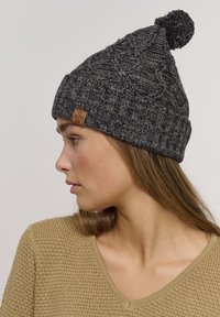 Beanie lavorato a maglia di colore grigio scuro con un motivo strutturato, caratterizzato da un pompon sulla sommità e un'etichetta marrone con logo sul lato.