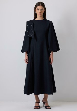 Abito nero con una silhouette fluida, maniche a campana lunghe e un dettaglio sulle spalle drappeggiato decorato con borchie. Indossato con tacchi neri con cinturini.