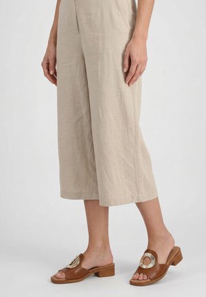 Persoon draagt beige wijde korte broek en bruine open teen sandalen met lage hak en een ronde metalen decoratie.