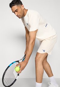 Light beige athletic outfit paired with tennis shorts. Holding a tennis racquet and ball, showcasing a subtle logo in blue.