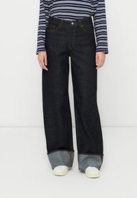 Mørkeblå vide jeans med høj talje, med væltede manchetter og kontrasterende kant. Kombinert med hvide sneakers og en stribet top.