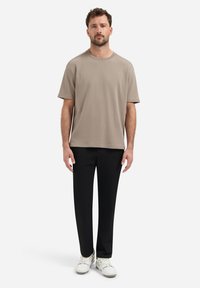 Beige t-shirt met korte mouwen in een relaxte pasvorm, gecombineerd met zwarte tapered broek en witte sneakers, staand voor een effen achtergrond.