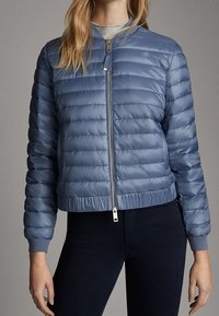 Femme portant une doudoune bleue matelassée, fermée, avec un pantalon noir et un t-shirt gris, debout devant un fond uni.