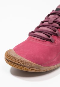 Zapatilla deportiva marrón con parte superior de suave ante, suela de goma texturizada y un sistema de cordones prominente. Punta redondeada con detalle de patrón de agarre.