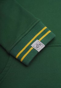 Grön sweatshirt med ribbade muddar som har två gula ränder, en vävd etikett på mudden och en slät yta.