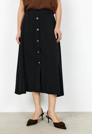 Žena v čiernej midi sukni na gombíky, hnedom pletenom topi a hnedých špicatých slingback podpätkoch stojaca na svetlej podlahe.