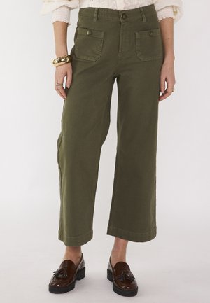 Donna che indossa pantaloni larghi cropped verde oliva con tasche frontali, mocassini marroni con nappine e una maglia bianca a maniche lunghe visibile ai polsi.