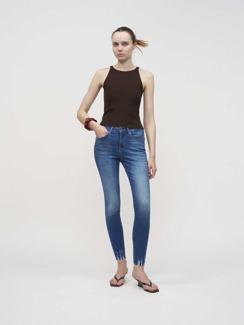 Jeune femme avec un haut marron sans manches, un jean skinny bleu avec des ourlets effilochés, des sandales à talons noires et un bracelet, posant devant un fond uni.