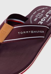 Tommy Hilfigeri plätud tumepunases värvuses, millel on tekstuuriga rihm, nahast aksent ja valged/punased triibud. Omab brändi logo jalatallal.