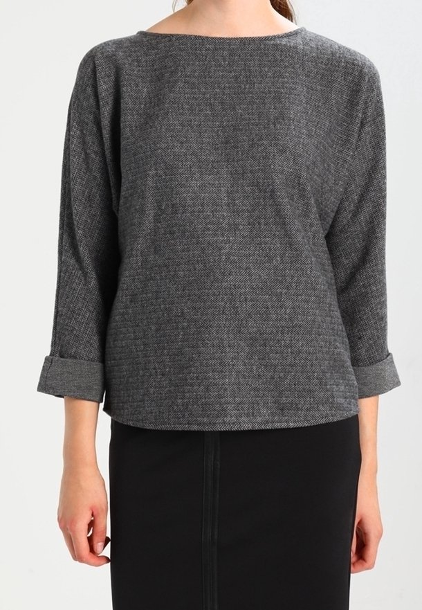 Femme portant un haut à manches longues texturé gris foncé avec les poignets retroussés et une jupe noire, debout devant un fond uni.