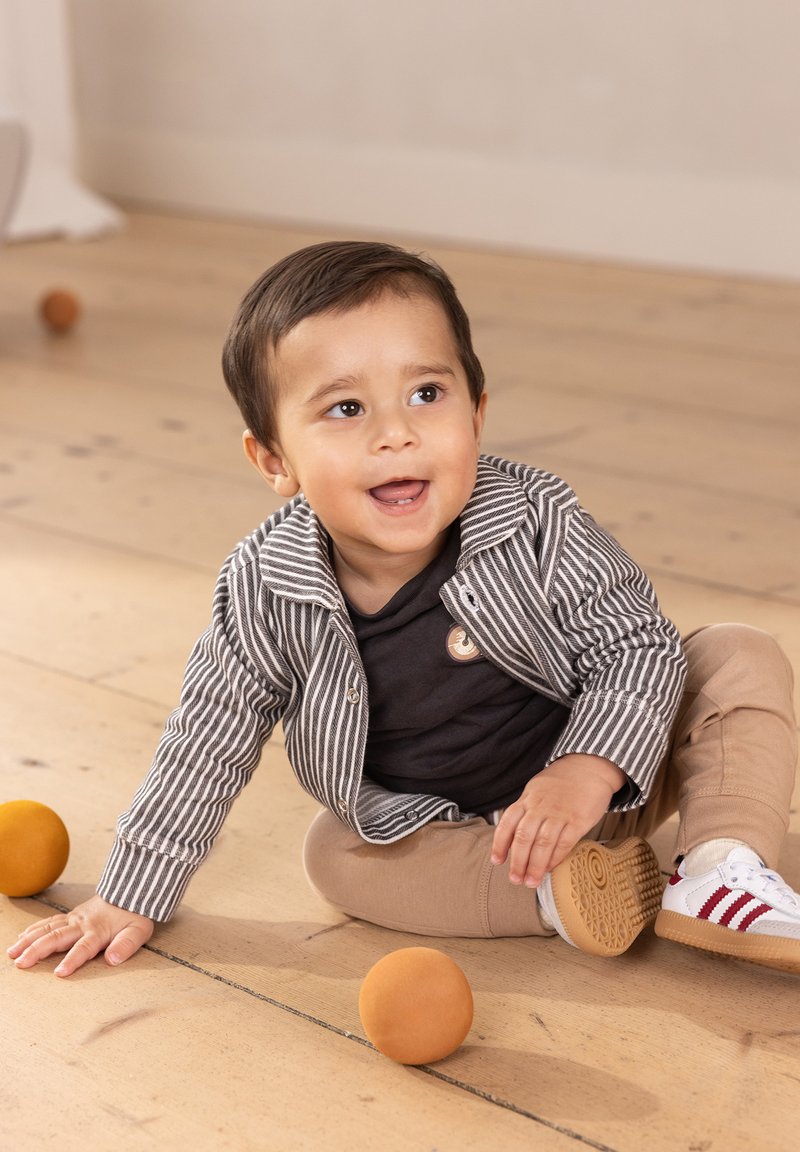 Gestreifte graue und weiße Knopfjacket, schwarzes Shirt, beige Hose und weiße Sneakers mit roten Streifen. Kind sitzt auf einem Holzboden mit orangen Bällen.