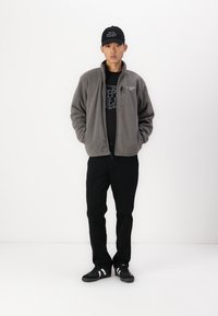 Grå fleecejacka, svart skjorta med grafik, svarta byxor och svarta sneakers med vita ränder. Enkel keps med text.