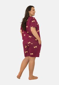 Kobieta z długimi brązowymi włosami ubrana w bordową bluzkę z krótkim rękawem i szorty z żółtymi wzorami w kokardy, stojąca boso bokiem.