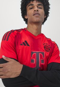 Rød fotballdrakt med svarte detaljer. Har v-hals, laglogo og skrå mønster. Modellen krysser armene, iført svarte langermede skjorter under.