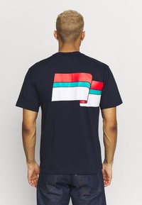 Mörkblå t-shirt med en djärv, flerfärgad grafik på baksidan; har röda, vita och akvamarin vågräta ränder. Kortsleeved.