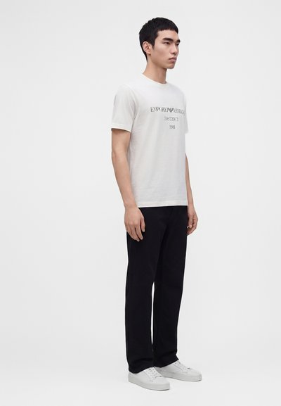 Νέος άνδρας που φοράει λευκό T-shirt με το λογότυπο Emporio Armani, μαύρο παντελόνι και λευκά αθλητικά παπούτσια μπροστά σε μονόχρωμο φόντο.
